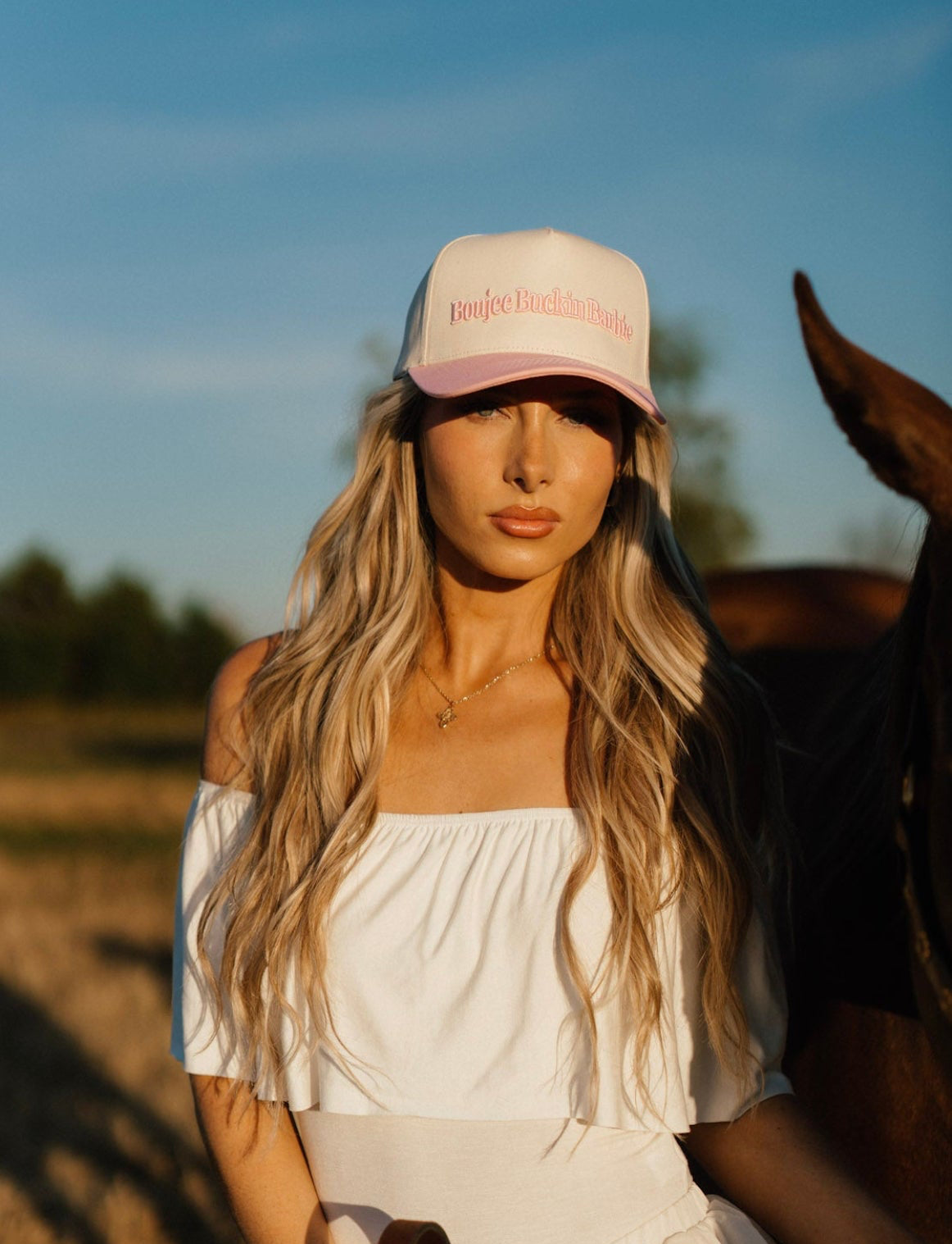Model wearing the Western Muse Boujee Buckin’ Barbie Hat with a white off-the-shoulder dress beside a horse.