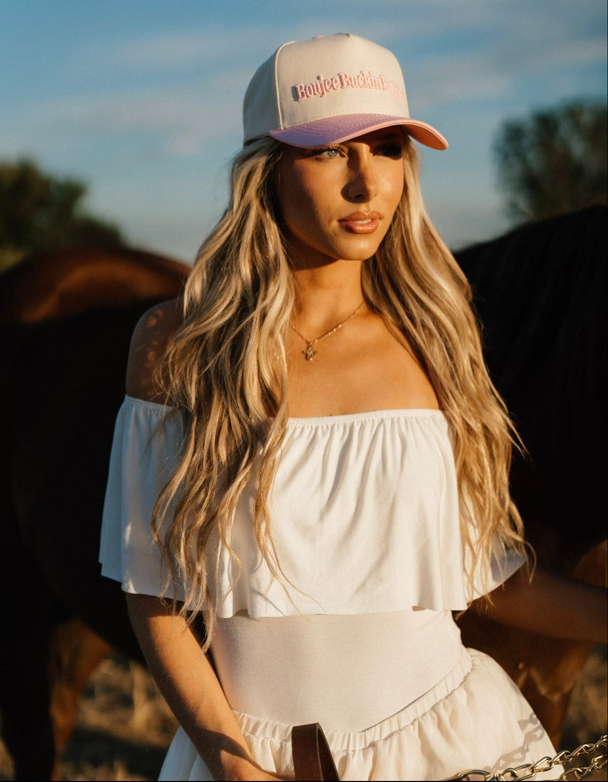 Model wearing the Western Muse Boujee Buckin’ Barbie Hat with a white off-the-shoulder dress beside a horse.